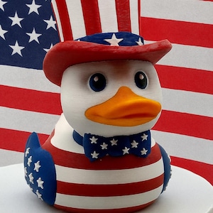 May include: A patriotic rubber ducky wearing a red, white, and blue Uncle Sam hat and bow tie. The duck features a white body with red and white striped accents and blue star details, set against an American flag backdrop.