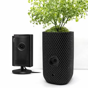 May include: A black rectangular security camera with the word cling printed on the side, positioned next to a black cylindrical planter with a diamond-patterned lattice exterior containing a small green leafy plant with a visible camera lens.