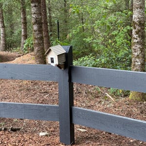 May include: A small, wooden birdhouse with a light brown roof and white siding is mounted on a black post of a gray wooden fence. The birdhouse is surrounded by green foliage and trees, with a forest background.