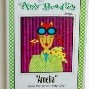 May include: A white card with a green dotted border features a cartoon cat named "Amelia" wearing blue goggles and a yellow polka dot scarf. The text "Amy Bradley Designs" is at the top.
