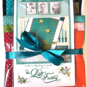 May include: A Simon Sea Turtle quilt pattern kit by The Quilt Factory, packaged in clear plastic with a teal ribbon. The pattern features photos of a baby and a finished quilt with fish and turtle designs. Includes assorted colorful fabric swatches.