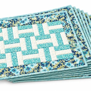 May include: A stack of rectangular fabric placemats featuring a central quilt-style pattern of alternating white and turquoise floral rectangles, surrounded by a dark blue border with a light blue and yellow floral print and a light blue edge.