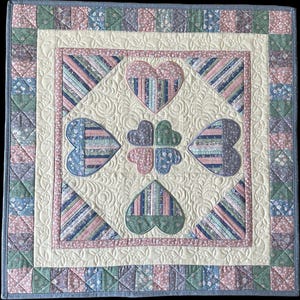 May include: A handmade quilt with a cream-colored center featuring heart and clover shapes in shades of pink, blue, and green. The quilt has a patterned border in coordinating colors and a quilted design throughout.