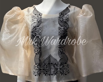 May include: A sheer, beige blouse with black embroidered floral designs. The blouse has puffed sleeves and a round neckline.