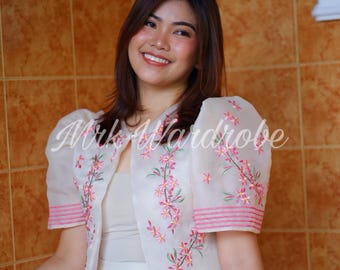 May include: A sheer, off-white bolero jacket with short, puffed sleeves. The jacket is embroidered with pink and green floral designs and horizontal pink stripes. The text "Mrk Wardrobe" is written across the front of the jacket.