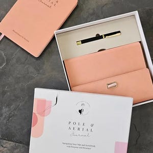 May include: A pink journal with the text "POLE & AERIAL Journal" on the cover. The journal is in a white box with a pink journal and a black pen.