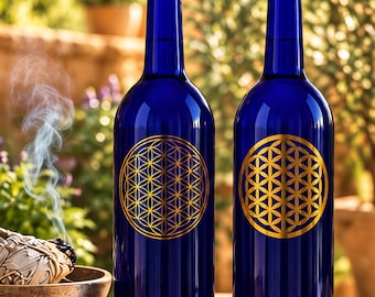 May include: Two tall, cylindrical cobalt blue glass bottles, each featuring a prominent, circular gold-coloured geometric flower of life pattern printed on the front surface. The bottles have a smooth, reflective finish and a narrow neck design.