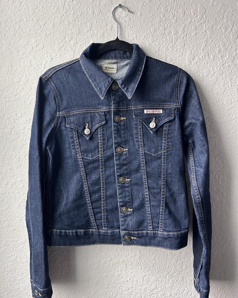 May include: A dark blue denim jacket featuring a pointed collar, long sleeves with buttoned cuffs, and a front button closure. It includes two chest pockets with buttoned flaps and a small rectangular label on the left pocket reading Hudson.