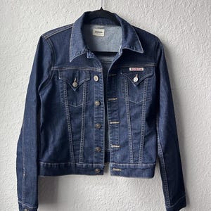 May include: A dark blue denim jacket featuring a pointed collar, long sleeves with buttoned cuffs, and a front button closure. The jacket includes two chest pockets with buttoned flaps and a small red brand label on the left pocket flap.
