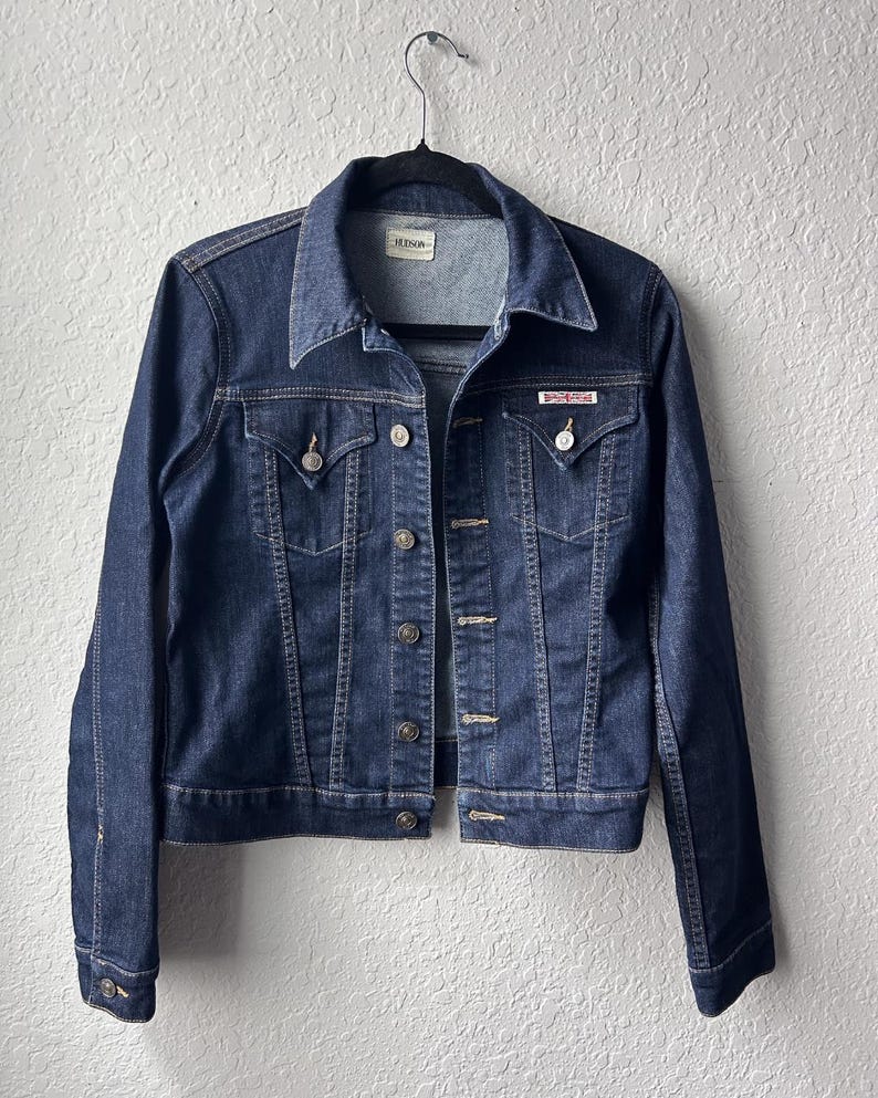 May include: A dark blue denim jacket featuring a pointed collar, long sleeves with buttoned cuffs, and a front button closure. The jacket includes two chest pockets with buttoned flaps and a small red brand label on the left pocket flap.
