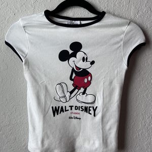 May include: White t-shirt with black trim featuring a graphic of Mickey Mouse in red shorts and black shoes. The text "WALT DISNEY STUDIOS" is printed below the character.