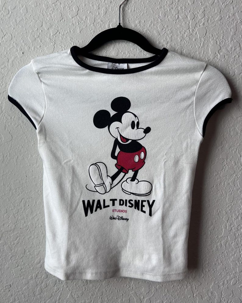 May include: White t-shirt with black trim featuring a graphic of Mickey Mouse in red shorts and black shoes. The text "WALT DISNEY STUDIOS" is printed below the character.