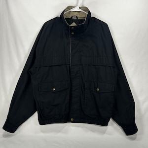 May include: A black jacket featuring a high collar with a tan interior lining, a front zipper closure with a snap-button placket, two large front pockets with snap-button flaps, and an elasticized waistband and cuffs for a secure fit.