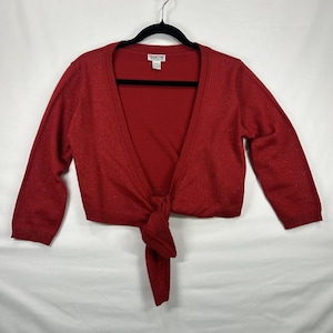 May include: A red knit shrug with three-quarter length sleeves and a tie-front closure. The garment features a subtle metallic shimmer throughout the fabric and includes a white interior label with the text Collection B.K.S. visible at the neckline.