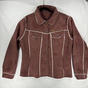 May include: A mauve-colored jacket with a soft, suede-like texture, featuring white faux-shearling trim along the seams, collar, and cuffs. The jacket includes a button-front closure, two chest pockets with button flaps, and long sleeves.