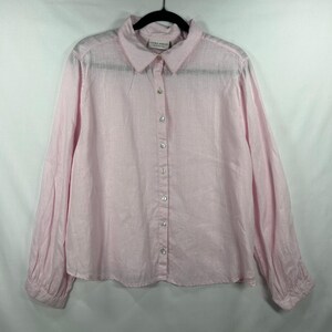 May include: A light pink long-sleeve button-down shirt with a classic collar and a front button placket. The shirt features a textured fabric finish and a label at the inner neckline that reads Laura Ashley. The sleeves have cuffs at the wrists.