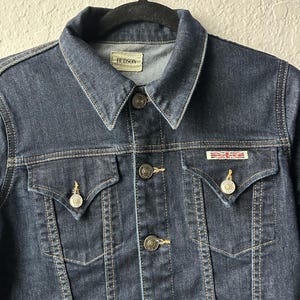 May include: A dark wash denim jacket featuring a pointed collar, a front button closure with metallic buttons, and two chest pockets with buttoned flaps. A small rectangular patch with a Union Jack flag design is attached above the right pocket.