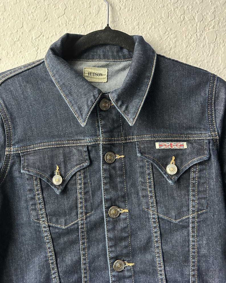 May include: A dark wash denim jacket featuring a pointed collar, a front button closure with metallic buttons, and two chest pockets with buttoned flaps. A small rectangular patch with a Union Jack flag design is attached above the right pocket.