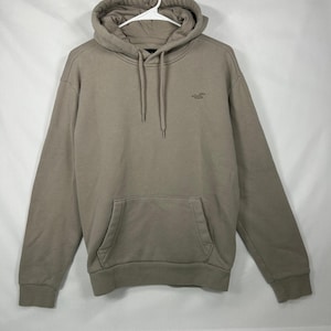 May include: A solid taupe-colored hooded sweatshirt featuring a front kangaroo pocket, long sleeves, and a drawstring hood. A small embroidered logo of a bird is visible on the upper left chest area. The garment is displayed on a white hanger.