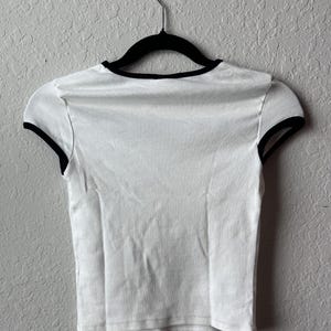 May include: White crop top with black trim around the neckline and sleeves, hanging on a black hanger. The shirt is made of a soft, lightweight fabric.
