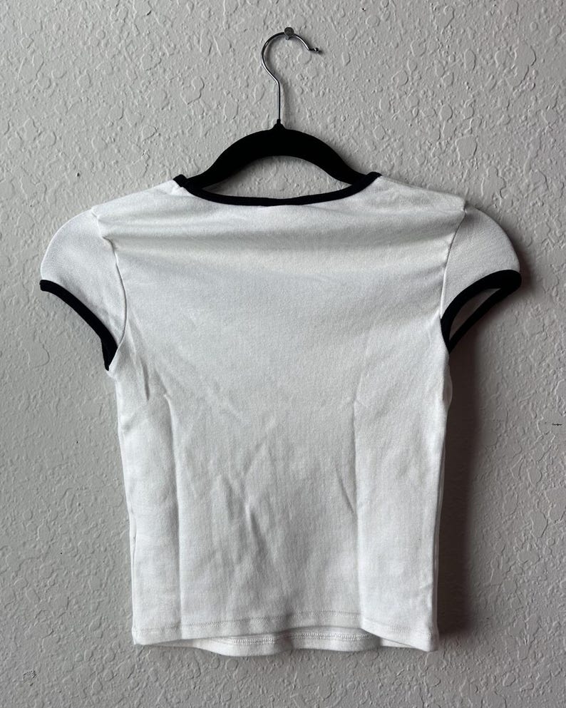 May include: White crop top with black trim around the neckline and sleeves, hanging on a black hanger. The shirt is made of a soft, lightweight fabric.