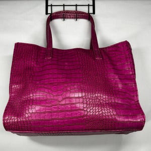 Puede incluir: Un bolso tote de color magenta vibrante con un patrón de textura de piel de cocodrilo en todo el exterior. El bolso incluye dos asas superiores resistentes unidas al borde superior, proporcionando una forma rectangular estructurada para llevar diversos objetos.