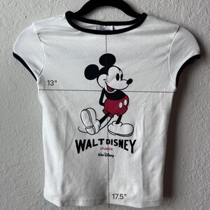 May include: White t-shirt with black trim featuring a Mickey Mouse graphic. The shirt has a 33 cm width and a 44.5 cm length. The Walt Disney Studios logo is printed below the character.