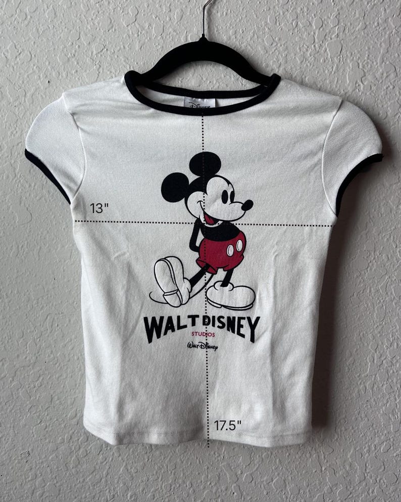 May include: White t-shirt with black trim featuring a Mickey Mouse graphic. The shirt has a 33 cm width and a 44.5 cm length. The Walt Disney Studios logo is printed below the character.