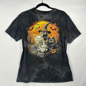 May include: A black tie-dye t-shirt featuring a graphic of three cats, one wearing a witch hat, set against a large yellow moon, bare trees, and flying bats. The scene includes orange pumpkins on the ground, creating a Halloween-themed design.