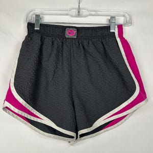 May include: Dark grey athletic shorts with a textured, crinkled fabric finish. The shorts feature bright pink side panels and white trim along the curved hem. A small rectangular patch at the center waistband displays a pink Nike swoosh logo.