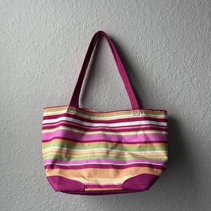 Puede incluir: Un bolso tote con rayas horizontales en tonos magenta, verde lima, naranja y blanco. El bolso tiene dos asas de hombro de color magenta y un panel base sólido del mismo color. La tela presenta una textura tejida visible en la superficie.