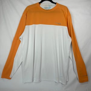 May include: A long-sleeve crew neck shirt featuring a colour-blocked design with an orange upper section and sleeves, and a white lower body. The Columbia PFG logo is printed in small text on the upper left chest area of the orange section.