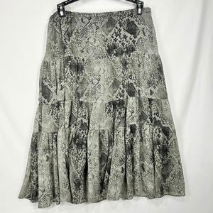 Peut inclure: Une jupe midi à étages avec un motif peau de serpent gris et noir. La jupe présente une taille élastique froncée et une silhouette fluide composée de trois étages horizontaux distincts qui créent une forme ample et volumineuse sur l'ensemble.
