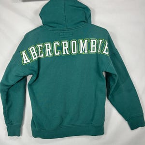 May include: A teal long-sleeve hooded sweatshirt featuring the word ABERCROMBIE printed in large white block letters with a lime green outline across the upper back. The garment includes a hood, long sleeves, and a ribbed hem and cuffs.
