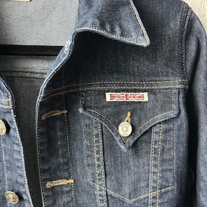 May include: A dark blue denim jacket featuring a pointed collar, gold-tone metal buttons, and a chest pocket with a flap. A rectangular patch with the text HUDSON and a Union Jack flag design is attached above the pocket. The jacket has visible stitching.
