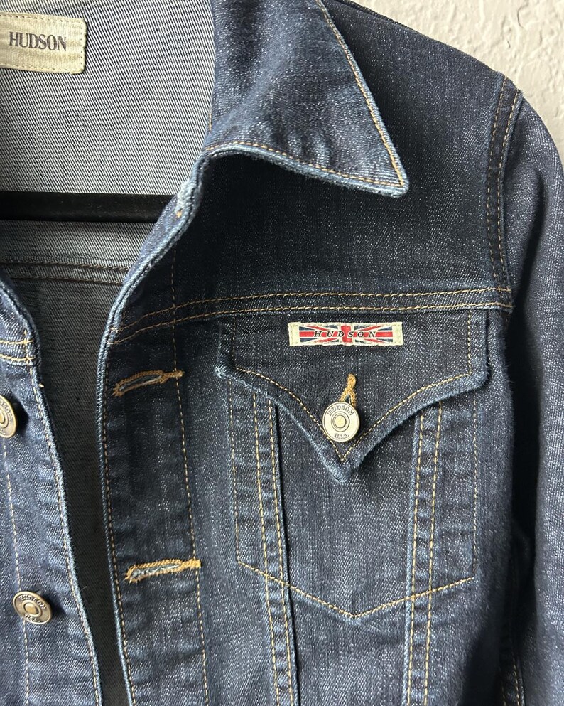 May include: A dark blue denim jacket featuring a pointed collar, gold-tone metal buttons, and a chest pocket with a flap. A rectangular patch with the text HUDSON and a Union Jack flag design is attached above the pocket. The jacket has visible stitching.