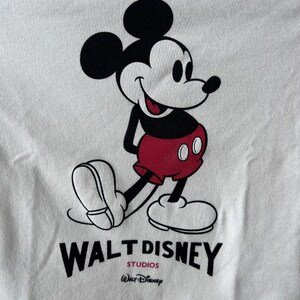 May include: White t-shirt with a smiling Mickey Mouse graphic. Mickey is in black, red, and white, with the Walt Disney Studios logo printed below. A classic and recognisable design.