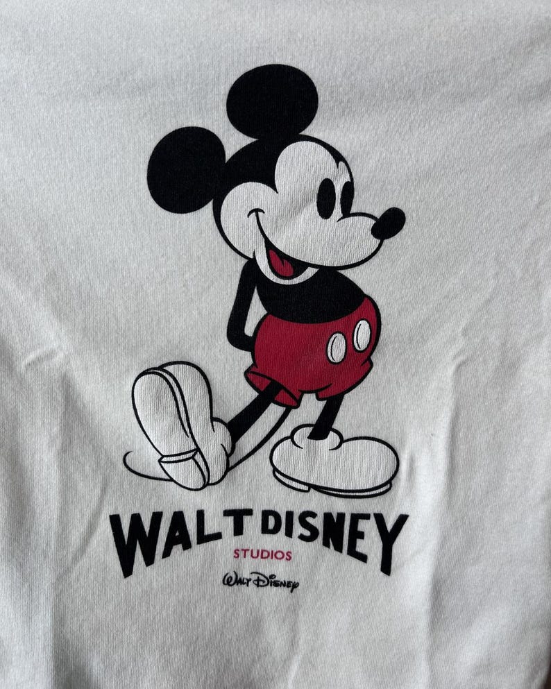 May include: White t-shirt with a smiling Mickey Mouse graphic. Mickey is in black, red, and white, with the Walt Disney Studios logo printed below. A classic and recognisable design.