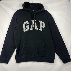 以下が含まれることがあります： 胸元に大きなライトグレーのブロック体でGAPの文字がプリントされた、長袖の黒いフード付きスウェットシャツ。前面にはカンガルーポケットがあり、黒い紐付きのフードとリブ編みの袖口が特徴です。