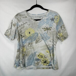 May include: A short-sleeve V-neck t-shirt featuring a light-colored background with a multi-colored print of yellow flowers, blue leaves, dragonflies, and cursive script text. The garment is displayed on a black hanger against a white background.