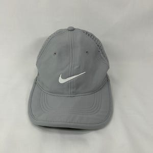 May include: A light gray baseball cap featuring a white Nike swoosh logo on the front panel. The cap is constructed with a smooth, lightweight fabric and includes perforated side panels for ventilation, a structured crown, and a curved brim.