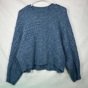 May include: A blue long-sleeve sweater featuring a textured cable knit pattern across the front and sleeves. The garment has a round ribbed neckline, dropped shoulders, and a slightly cropped hemline, constructed with a thick, heathered yarn.