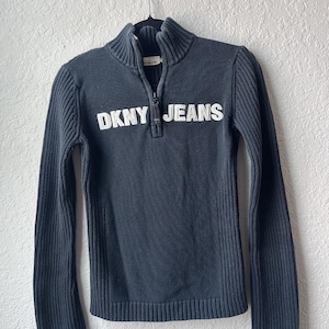 Könnte beinhalten: Ein dunkelgrauer DKNY JEANS Pullover mit gerippter Struktur und einem halben Reißverschluss. Der Markenname ist in großen weißen Buchstaben auf der Brust abgebildet. Der Pullover hängt an einem schwarzen Kleiderbügel.