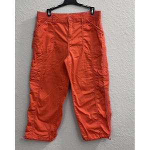May include: Bright orange cropped pants featuring a button and zipper fly closure, two front slash pockets, two side cargo pockets with flaps, and adjustable drawstring hems at the bottom of each leg, hanging from a black plastic hanger.