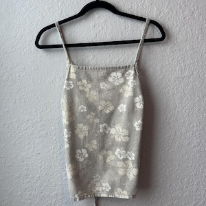 Op de afbeelding: Een lichtgrijze tanktop met een wit bloemenpatroon, hangend aan een zwarte hanger. De top heeft dunne bandjes en een vierkante halslijn. Het bloemmotief toont hibiscusbloemen.