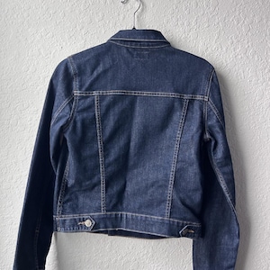May include: A dark blue denim jacket featuring a classic collar, long sleeves with buttoned cuffs, and a horizontal back yoke. The jacket includes vertical seam detailing, a bottom waistband with adjustable button tabs, and visible stitching.