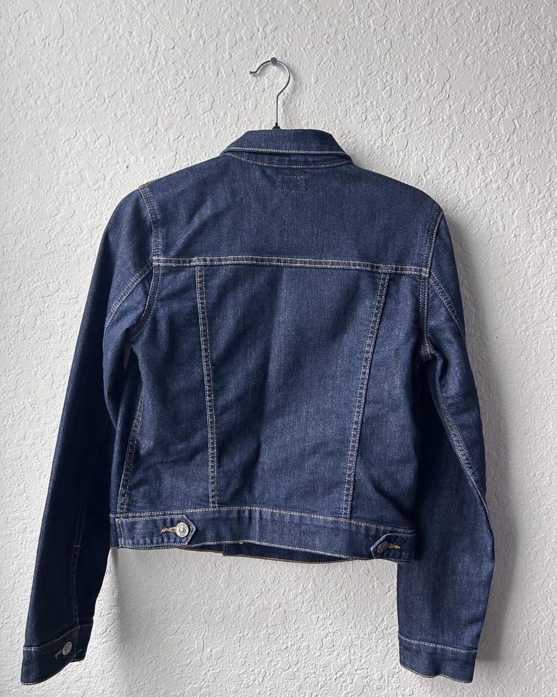 May include: A dark blue denim jacket featuring a classic collar, long sleeves with buttoned cuffs, and a horizontal back yoke. The jacket includes vertical seam detailing, a bottom waistband with adjustable button tabs, and visible stitching.