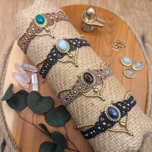 Puede incluir: Cuatro tobilleras de macramé con piedras preciosas de diferentes colores. Cada tobillera tiene un cierre de metal dorado y un dije colgante. Las tobilleras se muestran en una tela de arpillera marrón.
