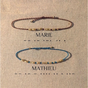 May include: Two friendship bracelets with gold beads and a brown and a blue cord. The bracelets are labeled "MARIE" and "MATHIEU" with morse code underneath.