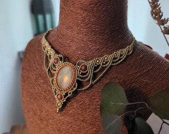 Puede incluir: Un collar de macramé verde y dorado con un gran colgante ovalado de piedra blanca. El collar está en un maniquí marrón.